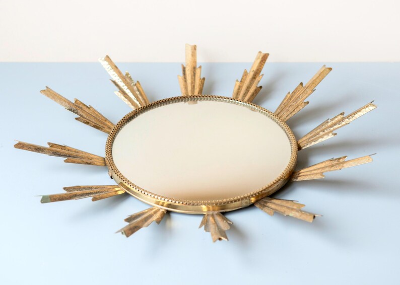 May include: A gold-coloured sunburst mirror with a round, reflective centre. The mirror's frame features radiating, stylised rays. The design is vintage-inspired, adding a decorative element to any space. The mirror is a decorative wall hanging.