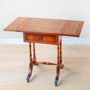 May include: A mahogany drop-leaf table with a leather top and a single drawer. The table has ornate legs with claw feet and casters. The table is approximately 76 cm long when fully extended.