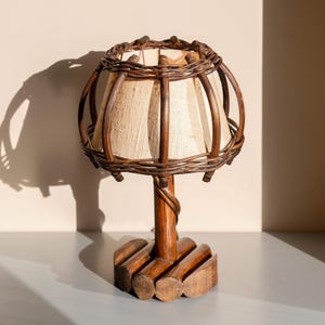 May include: A small, decorative table lamp with a woven wicker shade and base. The shade is made of light-colored fabric, and the frame is made of dark brown wicker. The base is made of wood and has a unique, rustic design.