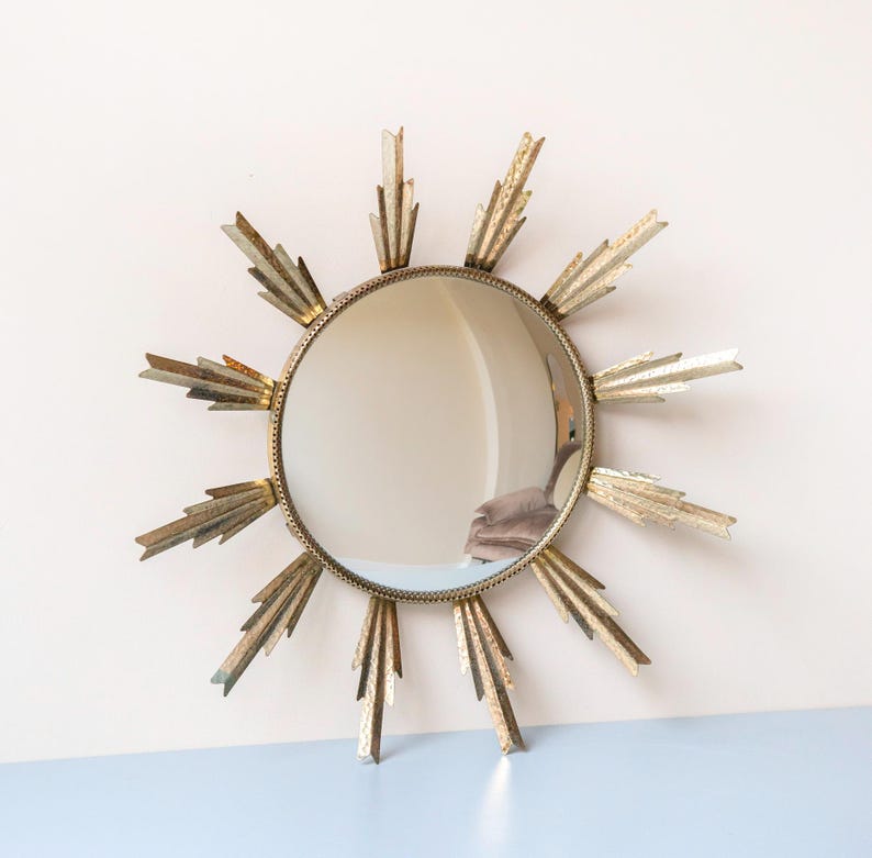 May include: A circular mirror with a gold-coloured sunburst frame. The mirror is round, with a decorative frame featuring textured, pointed rays. A stylish wall mirror, suitable for various interior styles.
