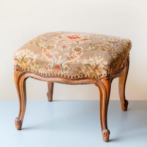 May include: A vintage footstool featuring a floral patterned cushion in tan, red, and green. The wooden frame is a warm brown with curved legs and decorative detailing. The cushion is secured with small tacks. The dimensions are approximately 40cm x 40cm.