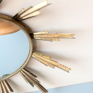 May include: A decorative sunburst mirror with a round, light blue mirror centre. The frame is gold-coloured with textured, pointed rays extending outwards. The mirror is a decorative wall hanging.