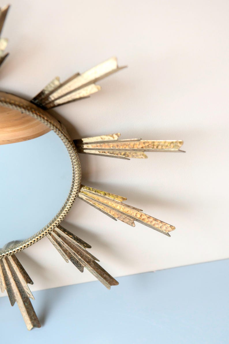 May include: A decorative sunburst mirror with a round, light blue mirror centre. The frame is gold-coloured with textured, pointed rays extending outwards. The mirror is a decorative wall hanging.