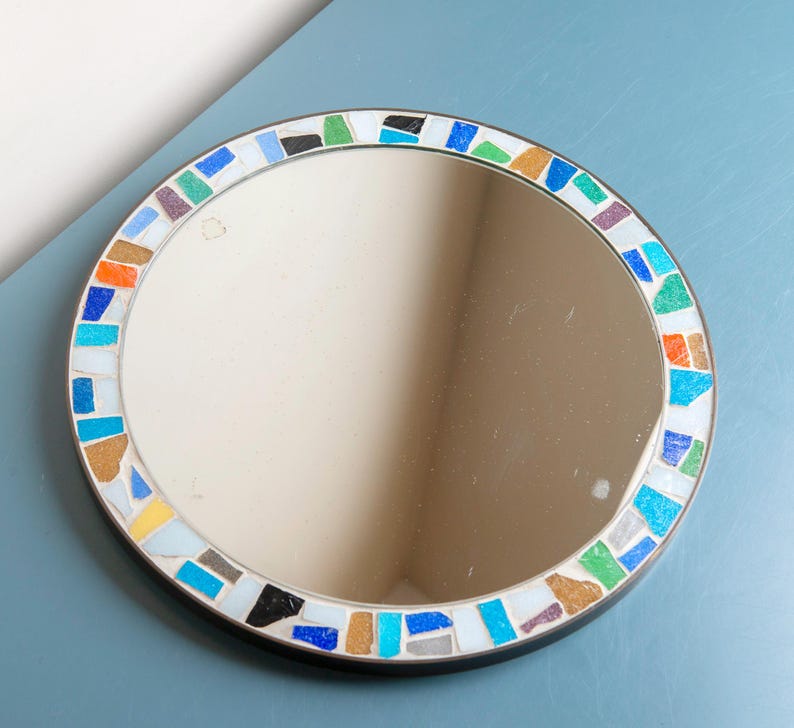May include: A round mirror with a colorful mosaic border. The border is made of small, irregular pieces of glass in shades of blue, green, orange, and white.
