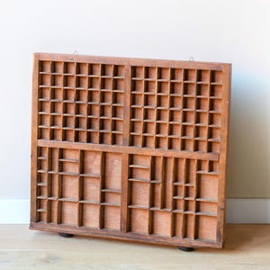 May include: A vintage wooden printer's tray with numerous small compartments. The tray is rectangular, made of light brown wood, and designed for organizing small items. It has a hanging hook on the top.