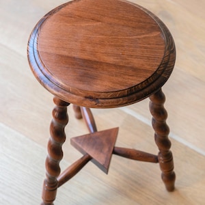May include: A round wooden stool with a smooth, polished brown surface and three legs featuring a twisted, spiral design. The legs are connected by a central triangular wooden brace that provides structural support to the base of the stool.