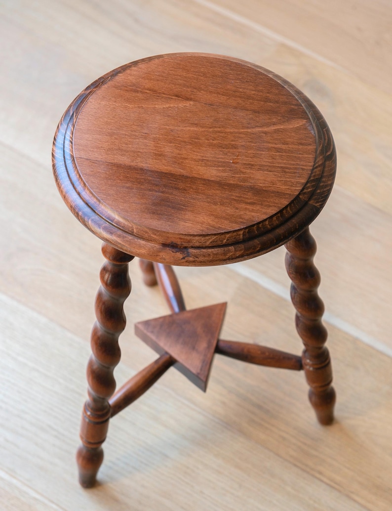 May include: A round wooden stool with a smooth, polished brown surface and three legs featuring a twisted, spiral design. The legs are connected by a central triangular wooden brace that provides structural support to the base of the stool.