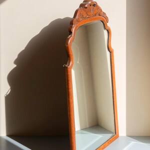 May include: A tall, arched wall mirror featuring a medium-toned wooden frame with a carved, decorative crest at the top. The mirror glass is clear and reflective, set within the smooth, contoured wooden border that follows the arched shape.