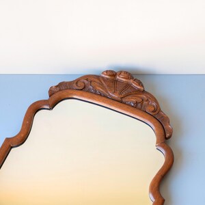 May include: A wall mirror with a medium-brown wooden frame featuring a scalloped, curved edge design. The top of the frame is adorned with a carved shell-like motif centred above the glass surface, which has a smooth, reflective finish.
