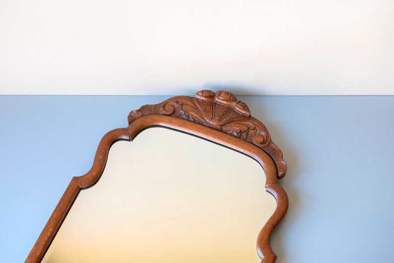 May include: A wall mirror with a medium-brown wooden frame featuring a scalloped, curved edge design. The top of the frame is adorned with a carved shell-like motif centred above the glass surface, which has a smooth, reflective finish.