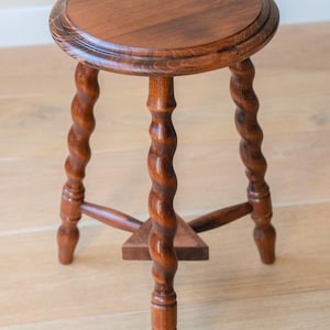 May include: A round wooden stool with a polished brown finish, featuring three legs with a distinct spiral twist design. The legs are connected by a small triangular support shelf near the base, providing stability to the circular seat top.
