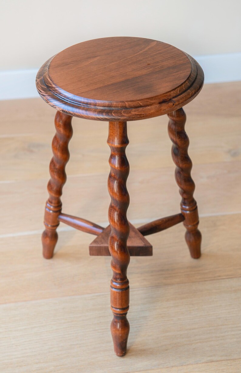 May include: A round wooden stool with a polished brown finish, featuring three legs with a distinct spiral twist design. The legs are connected by a small triangular support shelf near the base, providing stability to the circular seat top.