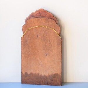 May include: A rectangular wooden mirror backing featuring a scalloped, arched top edge. The light brown wood grain is visible across the surface, and a thin yellow string is attached to two metal eyelets near the top for hanging purposes.