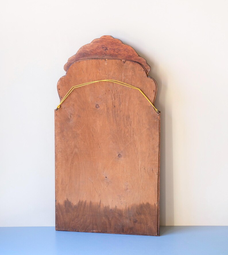 May include: A rectangular wooden mirror backing featuring a scalloped, arched top edge. The light brown wood grain is visible across the surface, and a thin yellow string is attached to two metal eyelets near the top for hanging purposes.