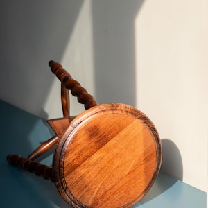 May include: A round wooden stool with a polished brown finish and a visible wood grain pattern. The stool features three legs with a distinct twisted, spiral-carved design and rounded feet, positioned at an angle on a flat surface.