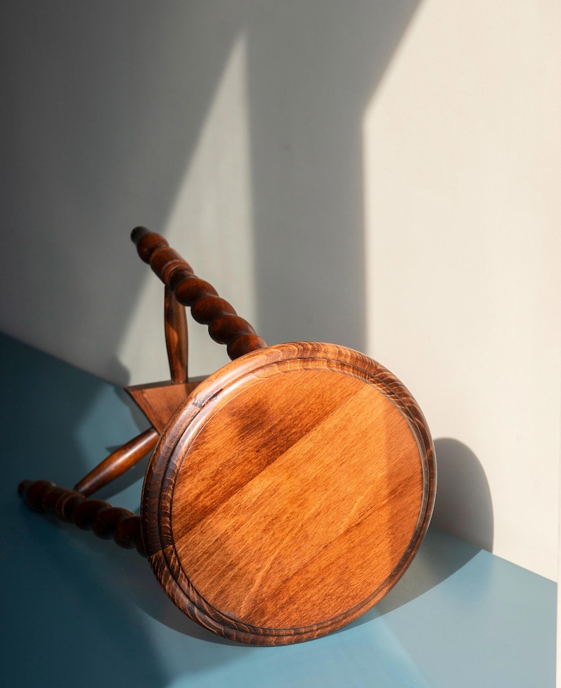 May include: A round wooden stool with a polished brown finish and a visible wood grain pattern. The stool features three legs with a distinct twisted, spiral-carved design and rounded feet, positioned at an angle on a flat surface.