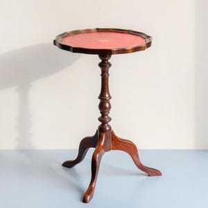 Op de afbeelding: Een vintage bijzettafel van donker hout met een rond, rood oppervlak. De tafel heeft een decoratieve, gedraaide centrale kolom en drie gebogen poten. De tafel is ongeveer 71 cm hoog.