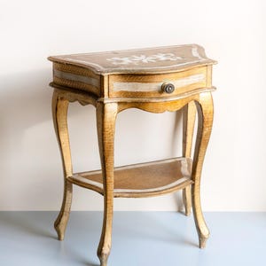 Puede incluir: Una pequeña mesa auxiliar antigua con forma de media luna. La mesa está pintada de dorado con un acabado envejecido y presenta un único cajón con un pomo redondo. La parte superior tiene un diseño floral y la mesa tiene un estante inferior. La mesa mide unos 71 cm de alto.
