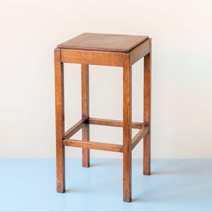 Puede incluir: Un taburete de madera con asiento cuadrado y cuatro patas. El taburete es de madera de tonos cálidos. El diseño es sencillo y funcional, con una barra de soporte horizontal cerca de la base de las patas. Altura aproximada 76 cm.