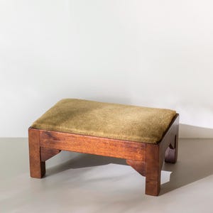 May include: A low rectangular footstool featuring a dark brown wooden frame with four sturdy legs and corner supports. The top surface is covered in a soft, textured olive-green fabric cushion that provides a flat, comfortable resting area.