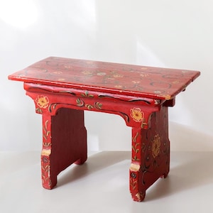 May include: A rectangular wooden stool painted in a vibrant red finish. The surface and legs are decorated with hand-painted floral motifs in yellow, white, and green. The stool features a flat top and two solid side panels with cut-out details.