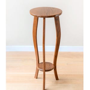 Puede incluir: Un soporte de madera para plantas con una parte superior redonda y un pequeño estante redondo. Tiene tres patas curvas y un soporte central. La madera tiene un tono marrón cálido. Altura aproximada 76 cm.