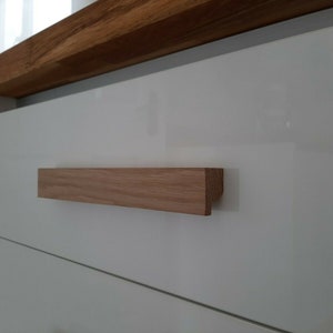 May include: A close-up of a white cabinet drawer with a wooden handle. The handle is rectangular and has a slightly rounded edge. The cabinet is made of a glossy white material.