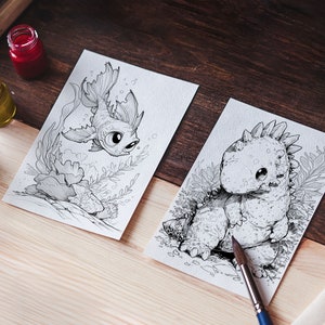 Op de afbeelding: Twee zwart-wit illustraties op wit papier, een van een vis en een van een dinosaurus. De illustraties liggen op een houten tafel met verfpotten en kleurpotloden.