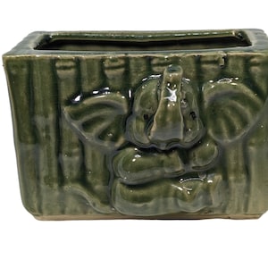 Puede incluir: Un macetero de cerámica verde con un diseño en relieve de un elefante. El macetero tiene una forma rectangular y un acabado liso y esmaltado.