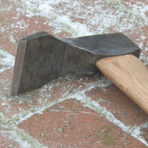 May include: Close-up of a forged steel axe head with a wooden handle. The axe head is dark gray and has a slightly curved blade. The wooden handle is light brown. The axe is resting on a red brick surface with patches of snow.