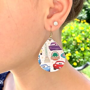 May include: A pair of white teardrop-shaped earrings with a Cars-themed print. The earrings feature images of Lightning McQueen, Mater, and other characters from the movie. The earrings are made of a white material with a gold-colored hook.