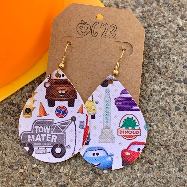 May include: A pair of white teardrop-shaped earrings with a Cars-themed print. The earrings feature images of Lightning McQueen, Mater, and other characters from the movie. The earrings are made of leather and have gold-colored hooks.