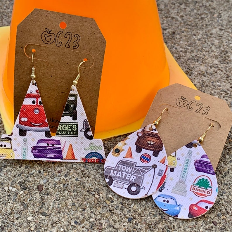 May include: A pair of dangle earrings with a white background and a Cars theme. The earrings are made of leather and feature images of Lightning McQueen, Mater, and other characters from the movie. The earrings are attached to gold-colored hooks.