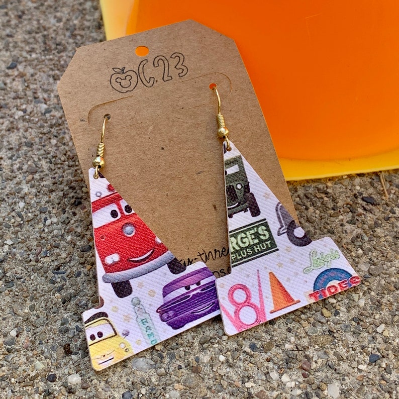 May include: A pair of dangle earrings featuring a white background with a colorful pattern of cars, trucks, and other vehicles. The earrings are shaped like a triangle with a rounded top and a flat bottom. The earrings are made of a lightweight material and have gold-colored hooks.