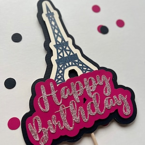 Puede incluir: Un adorno para pastel rosa y negro con una silueta de la Torre Eiffel y las palabras "Happy Birthday" en purpurina rosa.