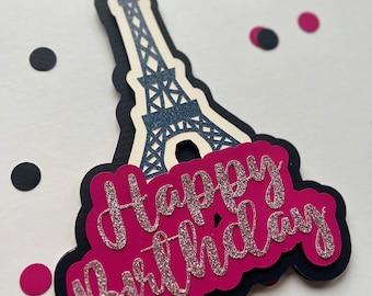May include: A pink and black cake topper with a silhouette of the Eiffel Tower and the words "Happy Birthday" in pink glitter.