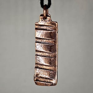 May include: A copper-toned rectangular pendant with horizontal lines and a textured surface, hanging from a black cord. The pendant has a loop at the top for the cord.