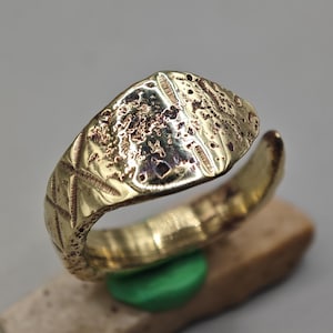May include: A gold-toned metal ring featuring a wide, textured, and pitted oval face. The band displays a cross-hatched, etched pattern along its sides. The ring has an open-ended design, showing a gap at the back of the band, with a metallic finish.