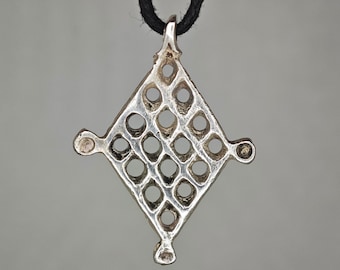 May include: A silver-toned metal pendant in a diamond shape with a lattice cutout pattern. The pendant features small circular protrusions at each of the four points and is suspended from a thin black cord, creating a geometric openwork design.