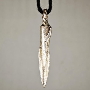 Può includere: Un ciondolo in metallo color argento a forma di punta di lancia o dente affusolato, con una superficie ruvida e usurata. Il ciondolo presenta un occhiello in metallo intrecciato sulla parte superiore, sospeso a una spessa corda nera.