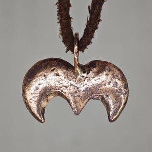 Könnte beinhalten: Ein bronzefarbener Anhänger mit einem Halbmond-Design, der an einer braunen Lederschnur hängt. Der Anhänger hat eine strukturierte Oberfläche und eine Schlaufe für die Schnur. Der Hintergrund ist einfarbig grau.