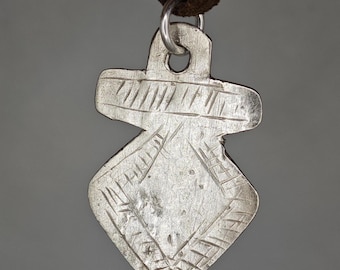 May include: A silver-toned metal pendant with a shield-like shape and a rectangular top section, featuring etched linear patterns along the edges and center. The pendant hangs from a dark brown leather cord through a circular silver jump ring.