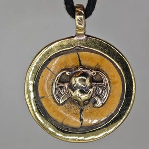 May include: A circular pendant featuring a central tan, cracked disc with a metallic gold-toned bat emblem. The disc is encased in a polished gold-toned metal frame with a matching bail at the top, suspended from a thick black cord necklace.