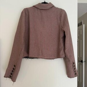 May include: A cropped blazer in a brown and black checkered pattern, hanging on a black hanger. The blazer has long sleeves with four buttons on each cuff and a frayed hemline. The jacket is displayed against a white wall.