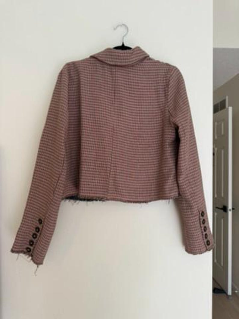 May include: A cropped blazer in a brown and black checkered pattern, hanging on a black hanger. The blazer has long sleeves with four buttons on each cuff and a frayed hemline. The jacket is displayed against a white wall.