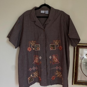 Può includere: Una camicia marrone a maniche corte con bottoni e ricami floreali e geometrici. La camicia ha un colletto e una chiusura con bottoni sul davanti.
