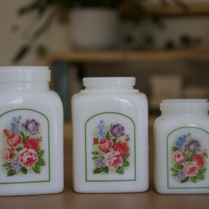 Op de afbeelding: Drie witte keramische potten met bloemenmotieven. De potten zijn in verschillende maten en hebben een groene rand rond de bloemenmotieven.