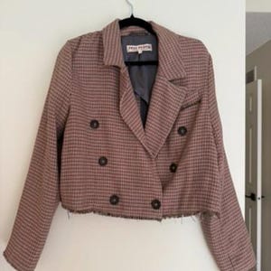 May include: A cropped, double-breasted blazer in a brown and black plaid pattern. The jacket has a notched collar, four dark brown buttons, and a frayed hem. The label reads "FREE PEOPLE".