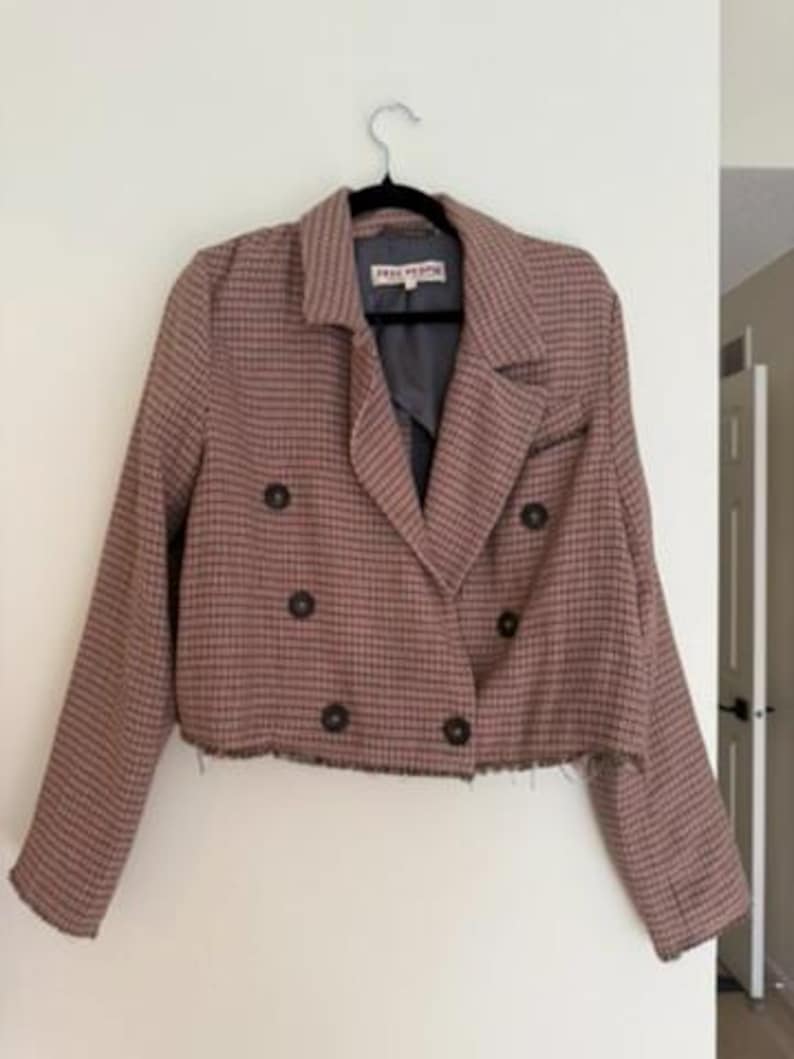 May include: A cropped, double-breasted blazer in a brown and black plaid pattern. The jacket has a notched collar, four dark brown buttons, and a frayed hem. The label reads "FREE PEOPLE".
