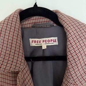 May include: Close-up of a coat with a red, white, and black plaid pattern. The coat has a gray lining and a label that reads "FREE PEOPLE". The coat is hanging on a black hanger.